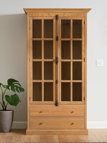 Tall cabinet with a washed grey finish, featuring a wooden frame, two glass-paneled doors, and two lower drawers. The cabinet is positioned against a light-colored wall, with a potted plant visible to the left. The overall design is elegant and showcases a blend of modern and rustic elements