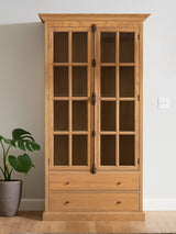 Tall cabinet with a washed grey finish, featuring a wooden frame, two glass-paneled doors, and two lower drawers. The cabinet is positioned against a light-colored wall, with a potted plant visible to the left. The overall design is elegant and showcases a blend of modern and rustic elements