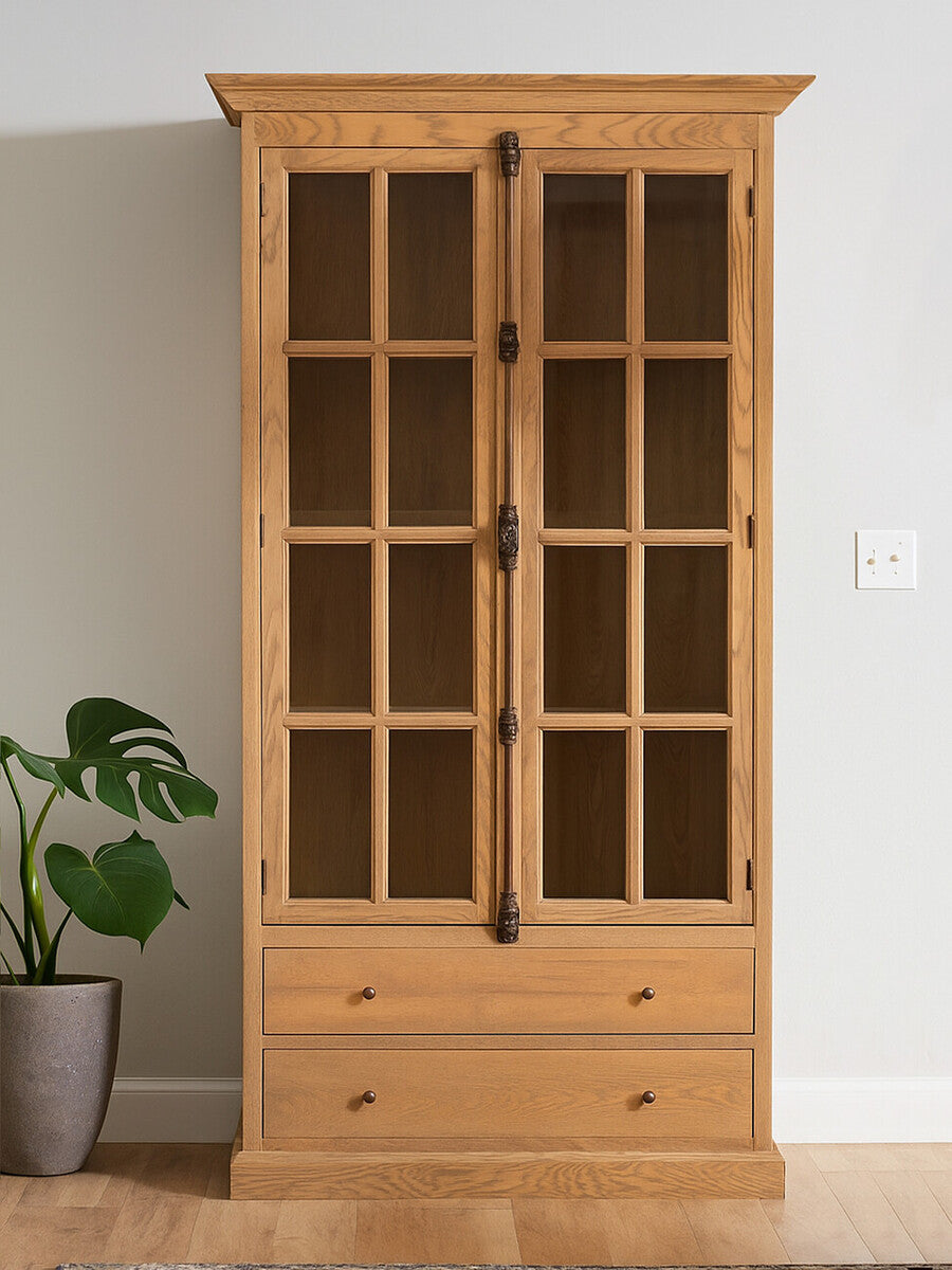 Tall cabinet with a washed grey finish, featuring a wooden frame, two glass-paneled doors, and two lower drawers. The cabinet is positioned against a light-colored wall, with a potted plant visible to the left. The overall design is elegant and showcases a blend of modern and rustic elements