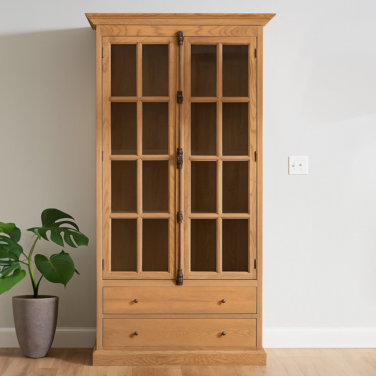 Tall cabinet with a washed grey finish, featuring glass-paneled doors and two lower drawers. The cabinet is made of oak wood and stands against a light-colored wall, with a potted plant beside it on the floor