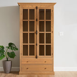 Tall cabinet with a washed grey finish, featuring glass-paneled doors and two lower drawers. The cabinet is made of oak wood and stands against a light-colored wall, with a potted plant beside it on the floor