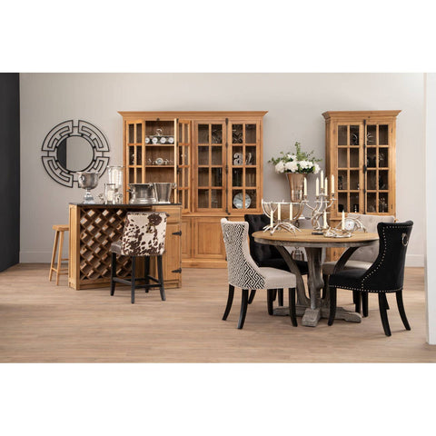 Large natural oak wood cabinet with glass doors, showcasing decorative items inside. The cabinet is positioned in a modern dining area featuring a round table with a metallic base and upholstered chairs in black and gray. A wine rack is visible beside the cabinet, and a wall clock adds to the decor. The floor is light wood, enhancing the warm tones of the furniture