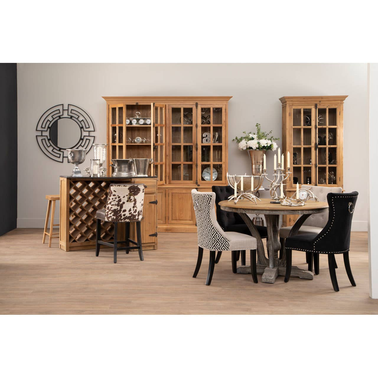 Large natural oak wood cabinet with glass doors, showcasing decorative items inside. The cabinet is positioned in a modern dining area featuring a round table with a metallic base and upholstered chairs in black and gray. A wine rack is visible beside the cabinet, and a wall clock adds to the decor. The floor is light wood, enhancing the warm tones of the furniture