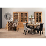 Large natural oak wood cabinet with glass doors, showcasing decorative items inside. The cabinet is positioned in a modern dining area featuring a round table with a metallic base and upholstered chairs in black and gray. A wine rack is visible beside the cabinet, and a wall clock adds to the decor. The floor is light wood, enhancing the warm tones of the furniture