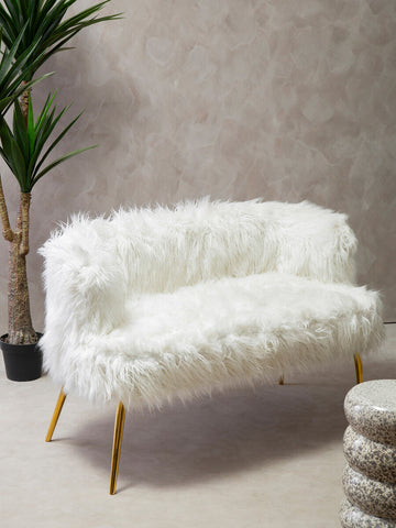 A plush white fur effect two-seater sofa with sleek gold legs, positioned next to a potted plant against a light-colored wall. The sofa features a curved backrest and a soft seat, creating a cozy and stylish seating option. A round, textured ottoman is visible in the foreground