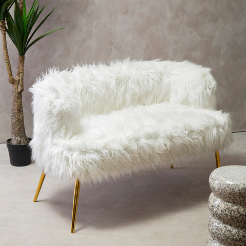 A plush white fur effect two-seater sofa with sleek gold legs, positioned against a neutral wall. A small potted plant is visible in the background, along with a round, textured ottoman nearby. The sofa's soft texture and modern design create a cozy and stylish seating area