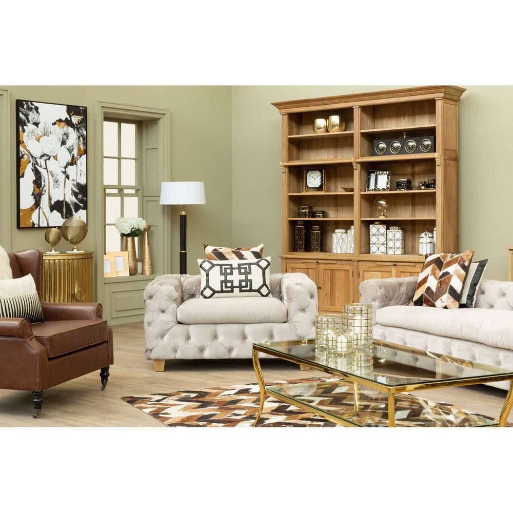 Beige velvet three-seat sofa with plush cushions, set in a cozy living room featuring a wooden bookshelf, a floor lamp, and decorative pillows on the sofa. The room has light green walls and a patterned area rug