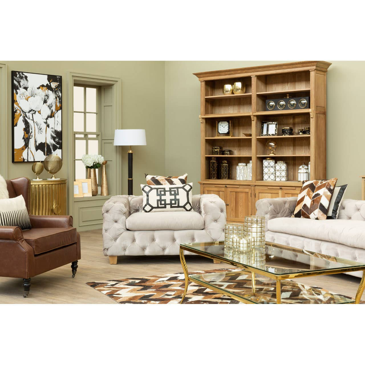 Beige velvet three-seat sofa with plush cushions, set in a cozy living room featuring a wooden bookshelf, a floor lamp, and decorative pillows on the sofa. The room has light green walls and a patterned area rug