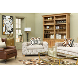 Beige velvet three-seat sofa with plush cushions, set in a cozy living room featuring a wooden bookshelf, a floor lamp, and decorative pillows on the sofa. The room has light green walls and a patterned area rug