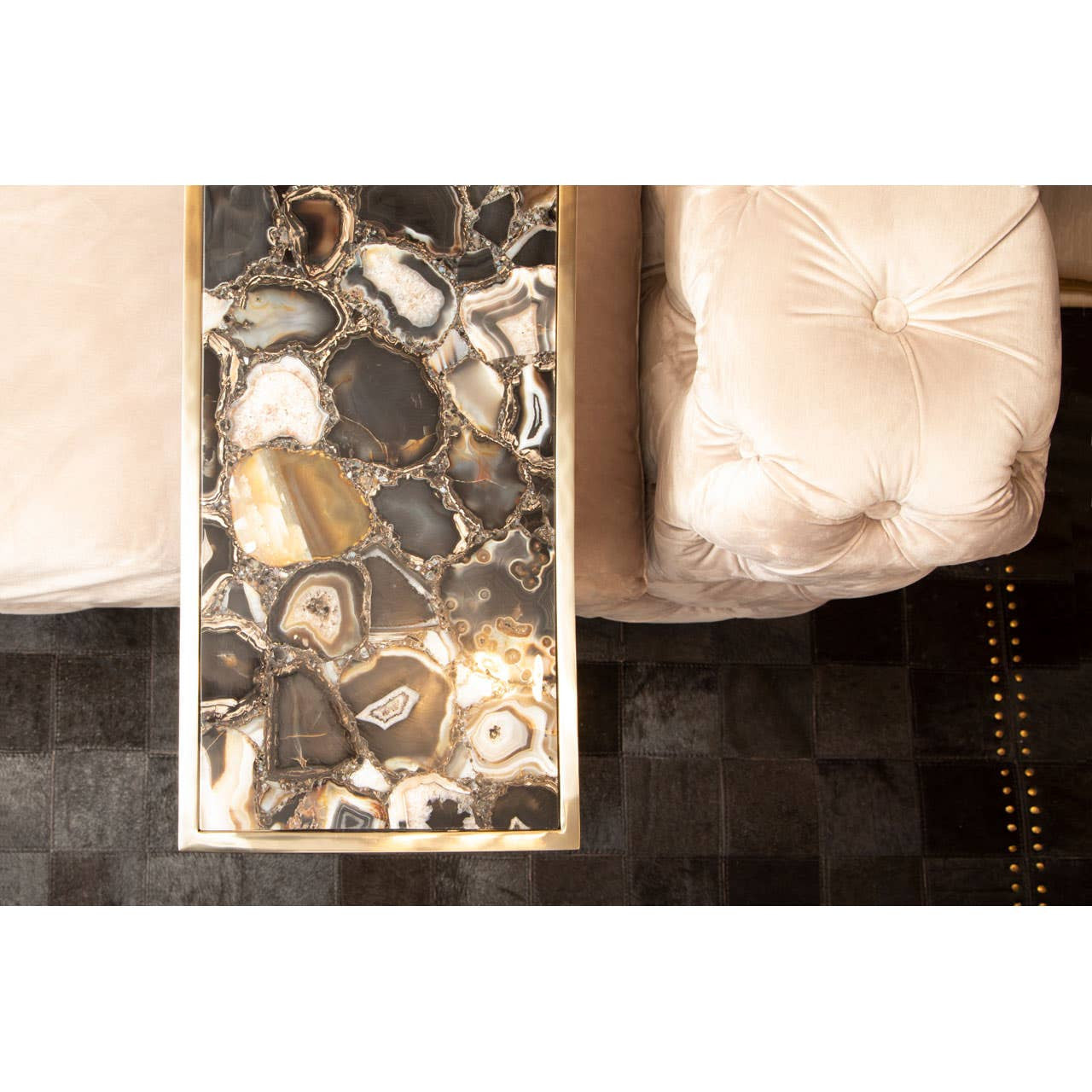 Beige velvet three-seat sofa with plush cushions, positioned next to a decorative coffee table featuring an abstract design in shades of brown and gold. The sofa's soft texture and elegant design complement the modern decor of the room