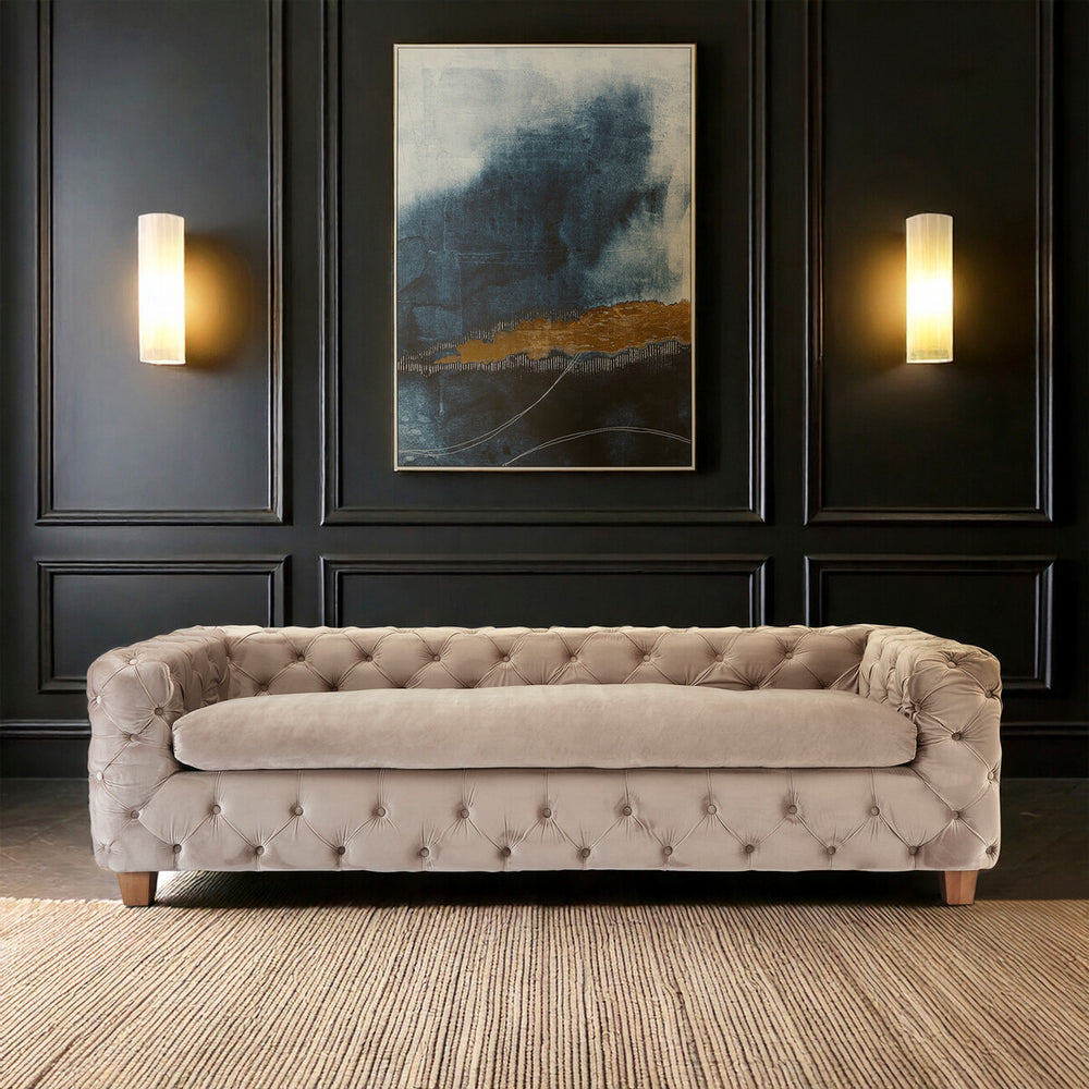 Beige velvet three-seat sofa with tufted upholstery, wooden legs, and a sleek design, positioned against a dark wall featuring a large abstract painting and two wall sconces. The floor is made of light-colored wood