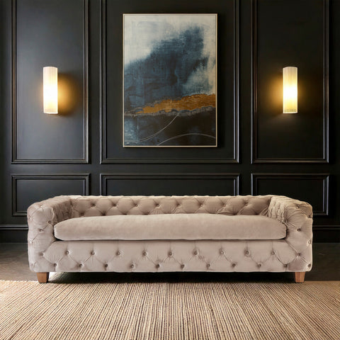 Beige velvet three-seat sofa with tufted upholstery, wooden legs, and a sleek design, positioned against a dark wall featuring a large abstract painting and two wall sconces. The floor is made of light-colored wood