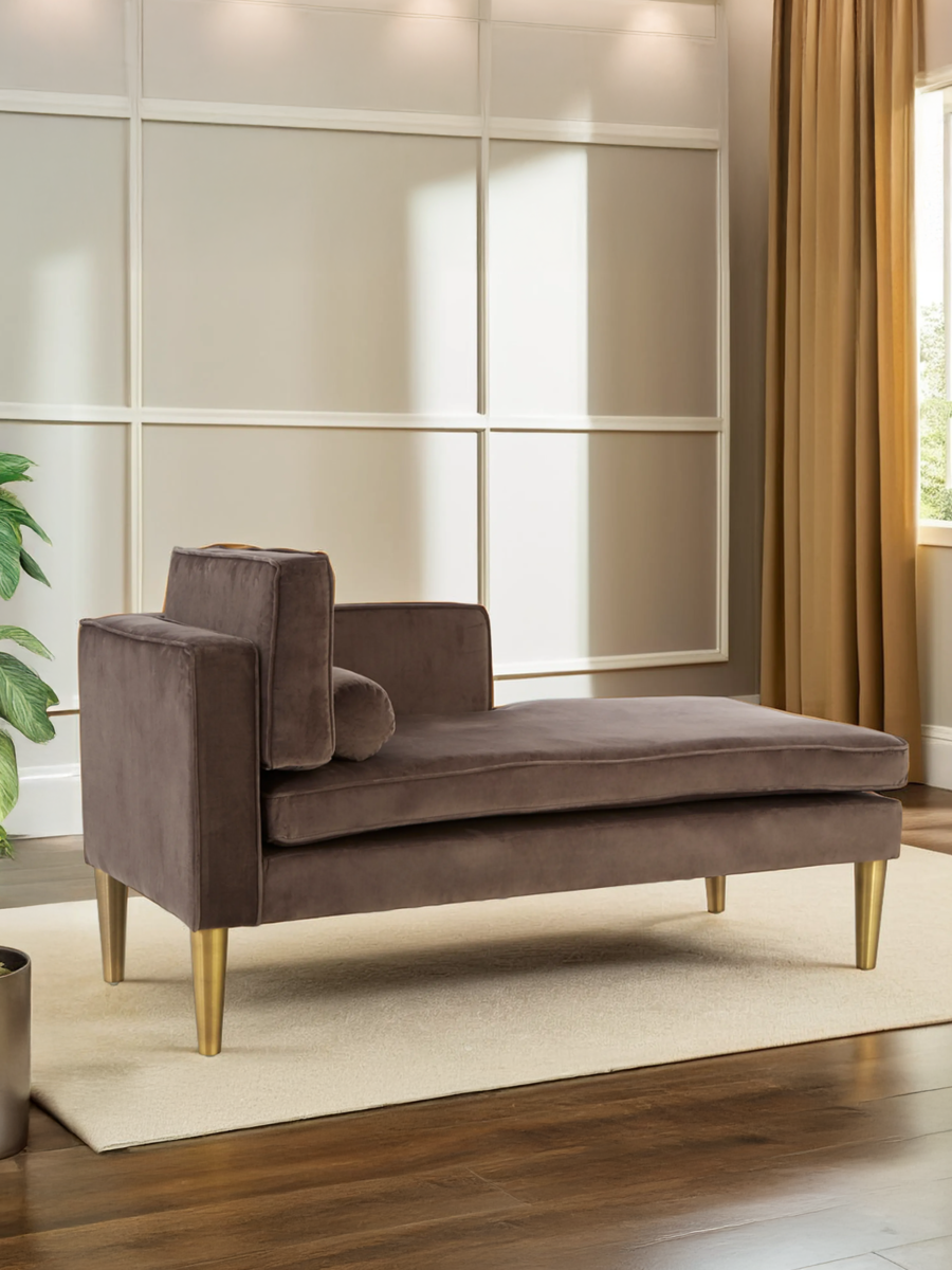 Grey velvet left-hand chaise longue with a sleek design, featuring a plush seat and a supportive backrest. The chaise is supported by wooden legs and includes a matching cushion, set against a backdrop of neutral walls and warm curtains. A potted plant is visible nearby, adding a touch of greenery to the scene