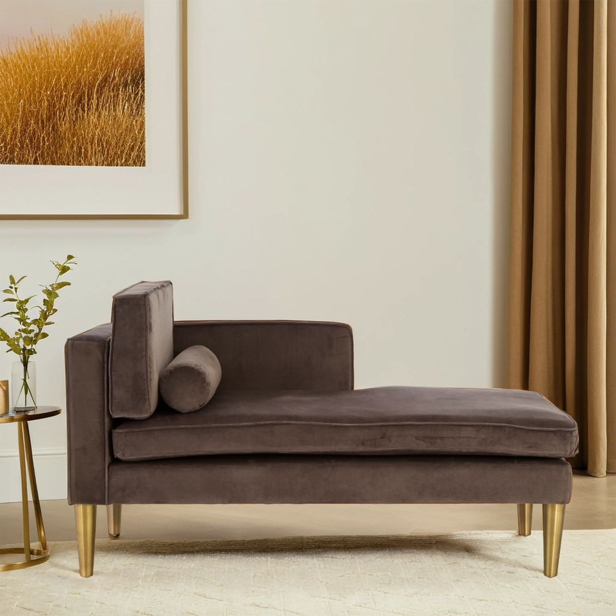 Grey velvet left-hand chaise longue with a sleek design, featuring a rounded backrest and a decorative bolster pillow. It is supported by elegant gold legs and positioned on a light-colored rug, with a backdrop of neutral walls and a framed artwork. A small side table with a vase of flowers is placed nearby