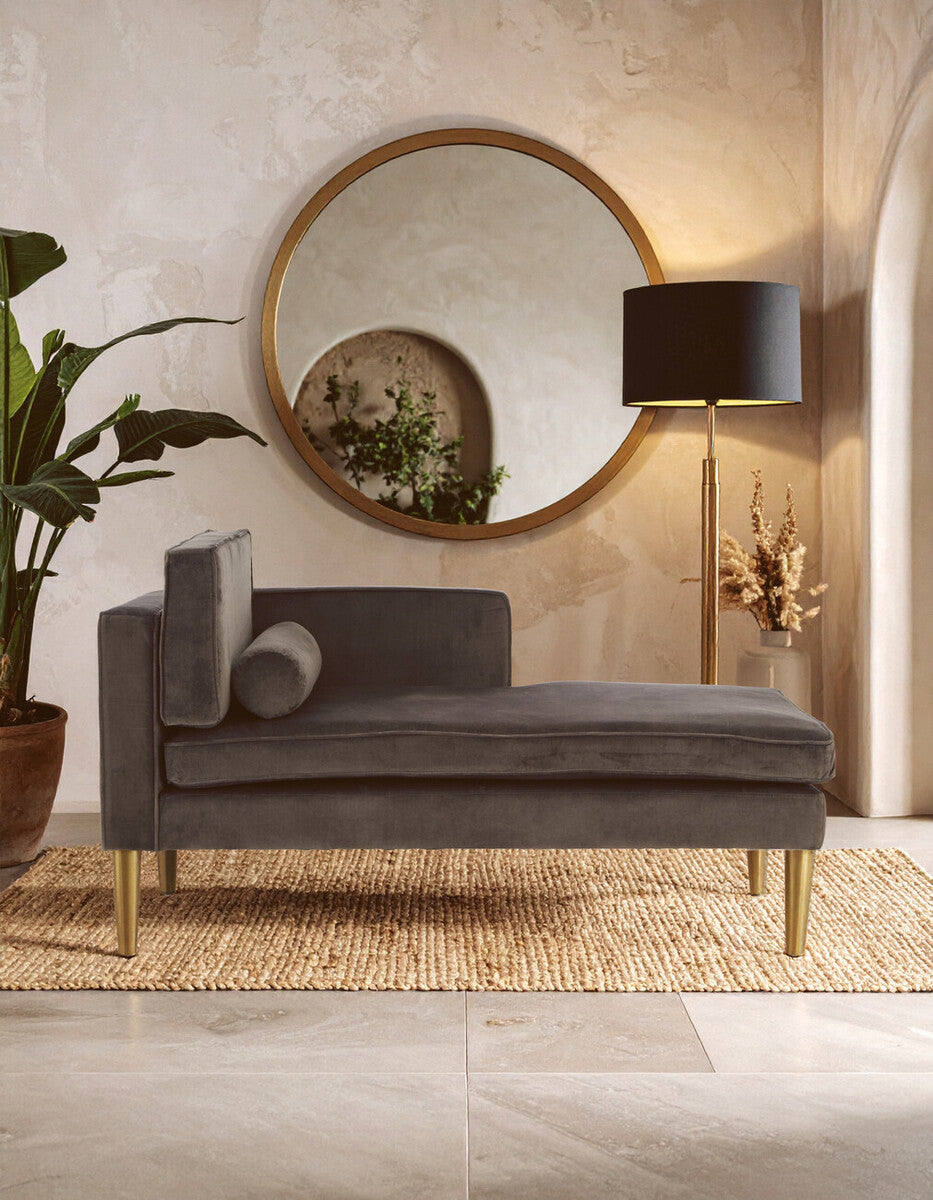 Grey velvet left-hand chaise longue with a curved backrest, a decorative cushion, and wooden legs, positioned in a stylish interior featuring a large round mirror, a floor lamp, and a potted plant. The setting has warm tones and a textured rug