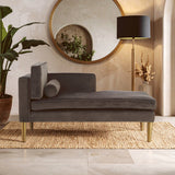 Grey velvet left-hand chaise longue with a sleek design, featuring a plush seat and a rounded backrest. A decorative cushion rests on the side, and the chaise is supported by elegant gold legs. The setting includes a large round mirror and a floor lamp, with a potted plant in the background, all placed on a textured rug