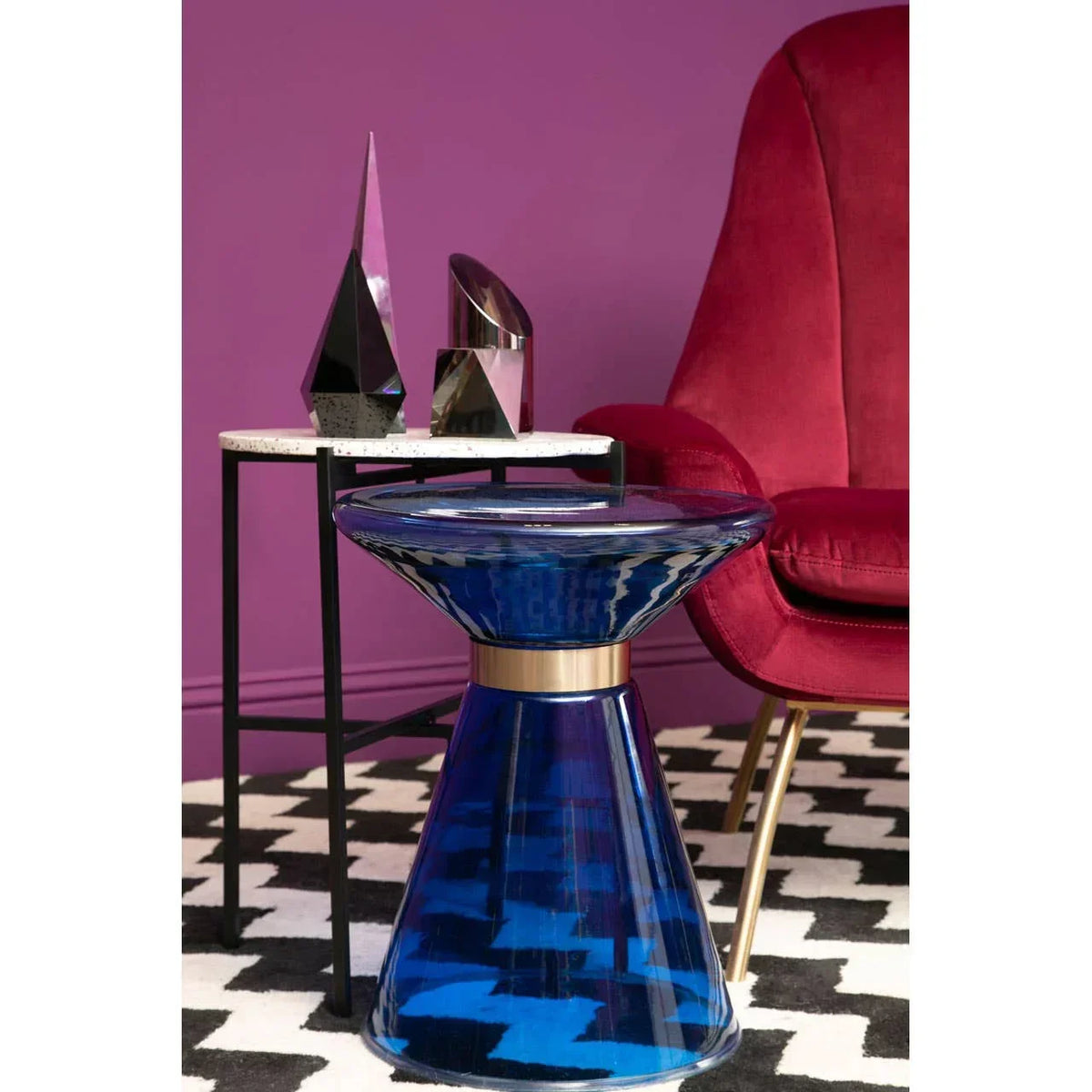Blue glass side table beside marble accent table and red velvet armchair on patterned rug