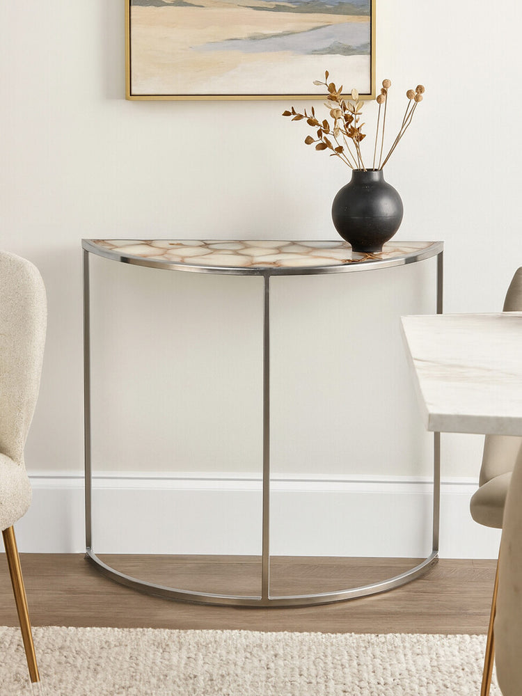 Half moon console table featuring a white agate top with natural patterns, supported by a sleek metal frame. The table is positioned against a light-colored wall, accompanied by a decorative vase and a small arrangement of dried flowers. A neutral-toned chair is visible to the left, and a portion of a dining table is seen to the right