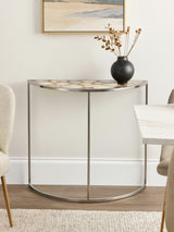 Half moon console table featuring a white agate top with natural patterns, supported by a sleek metal frame. The table is positioned against a light-colored wall, accompanied by a decorative vase and a small arrangement of dried flowers. A neutral-toned chair is visible to the left, and a portion of a dining table is seen to the right