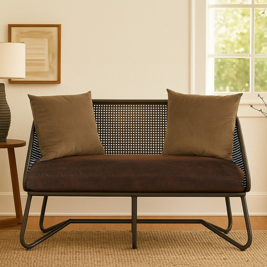 A two-seat sofa with a sleek design featuring curved legs, upholstered in a dark fabric. The sofa is adorned with two square, textured throw pillows in a complementary color. It is positioned in a cozy room with natural light coming through a window, and a light-colored rug is visible beneath it. A lamp and framed artwork are in the background, adding to the inviting atmosphere