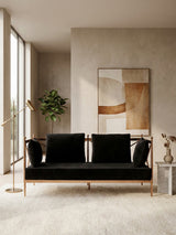 A stylish two-seater sofa featuring a black velvet upholstery and a rose gold metal frame, positioned in a modern living space with large windows, a potted plant, and a decorative vase on a light-colored rug. A large mirror is mounted on the wall behind the sofa, reflecting the room's contemporary design elements