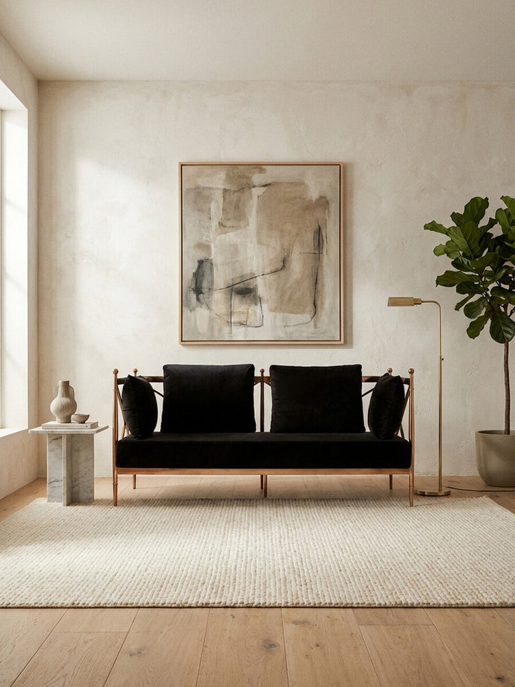 A stylish two-seater sofa upholstered in black velvet, featuring a sleek rose gold frame. The sofa is positioned in a bright room with a large window, a textured area rug beneath it, and a decorative plant nearby. A modern artwork hangs on the wall behind the sofa, adding a contemporary touch to the space