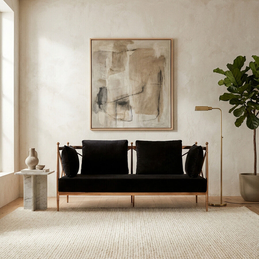 A stylish two-seater sofa upholstered in black velvet, featuring a sleek rose gold frame. The sofa is positioned in a bright room with a large window, a textured area rug beneath it, and a decorative plant nearby. A modern artwork hangs on the wall behind the sofa, adding a contemporary touch to the space