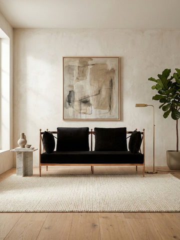 A stylish two-seater sofa upholstered in black velvet, featuring a sleek rose gold frame. The sofa is positioned in a bright room with a large window, a textured area rug beneath it, and a decorative plant nearby. A modern artwork hangs on the wall behind the sofa, adding a contemporary touch to the space