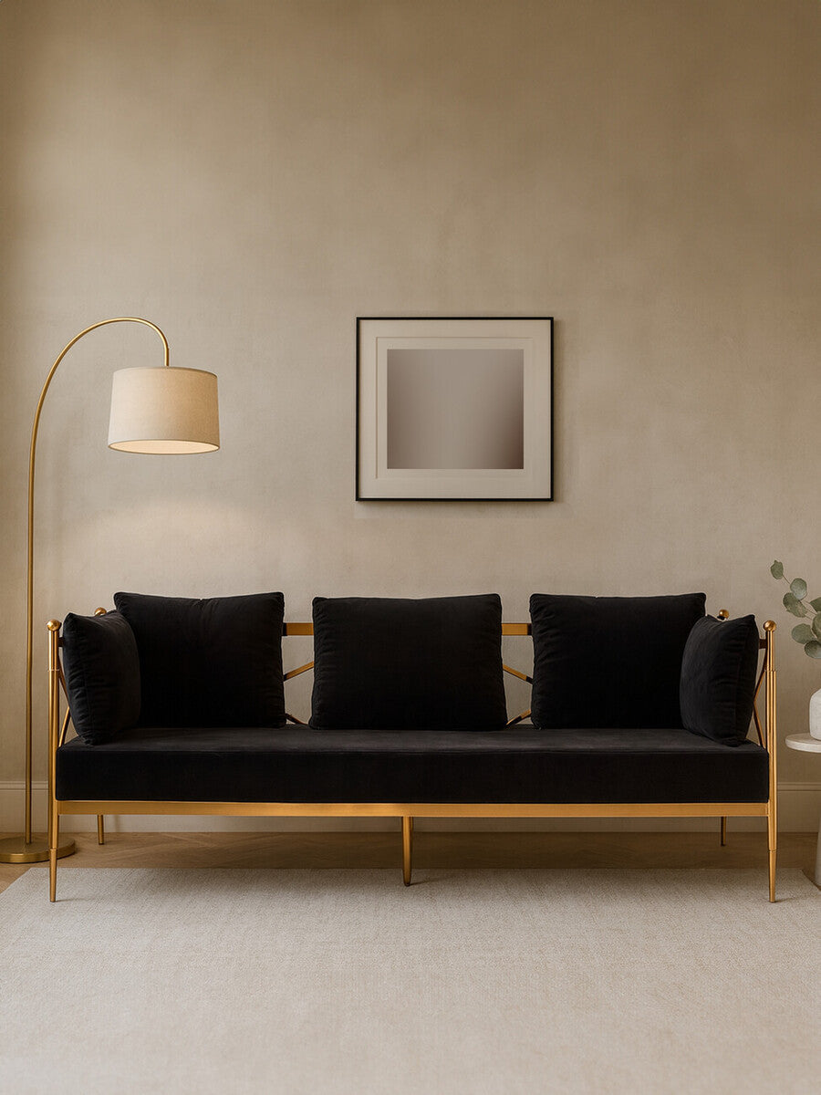 A three-seater sofa upholstered in black velvet, featuring a sleek rose gold frame. The sofa has three plush cushions and is positioned against a neutral-colored wall. A modern floor lamp stands to the left, and a minimalist artwork is displayed above the sofa. A small plant is visible on the right side of the image