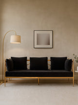 A three-seater sofa upholstered in black velvet, featuring a sleek rose gold frame. The sofa has three plush cushions and is positioned against a neutral-colored wall. A modern floor lamp stands to the left, and a minimalist artwork is displayed above the sofa. A small plant is visible on the right side of the image
