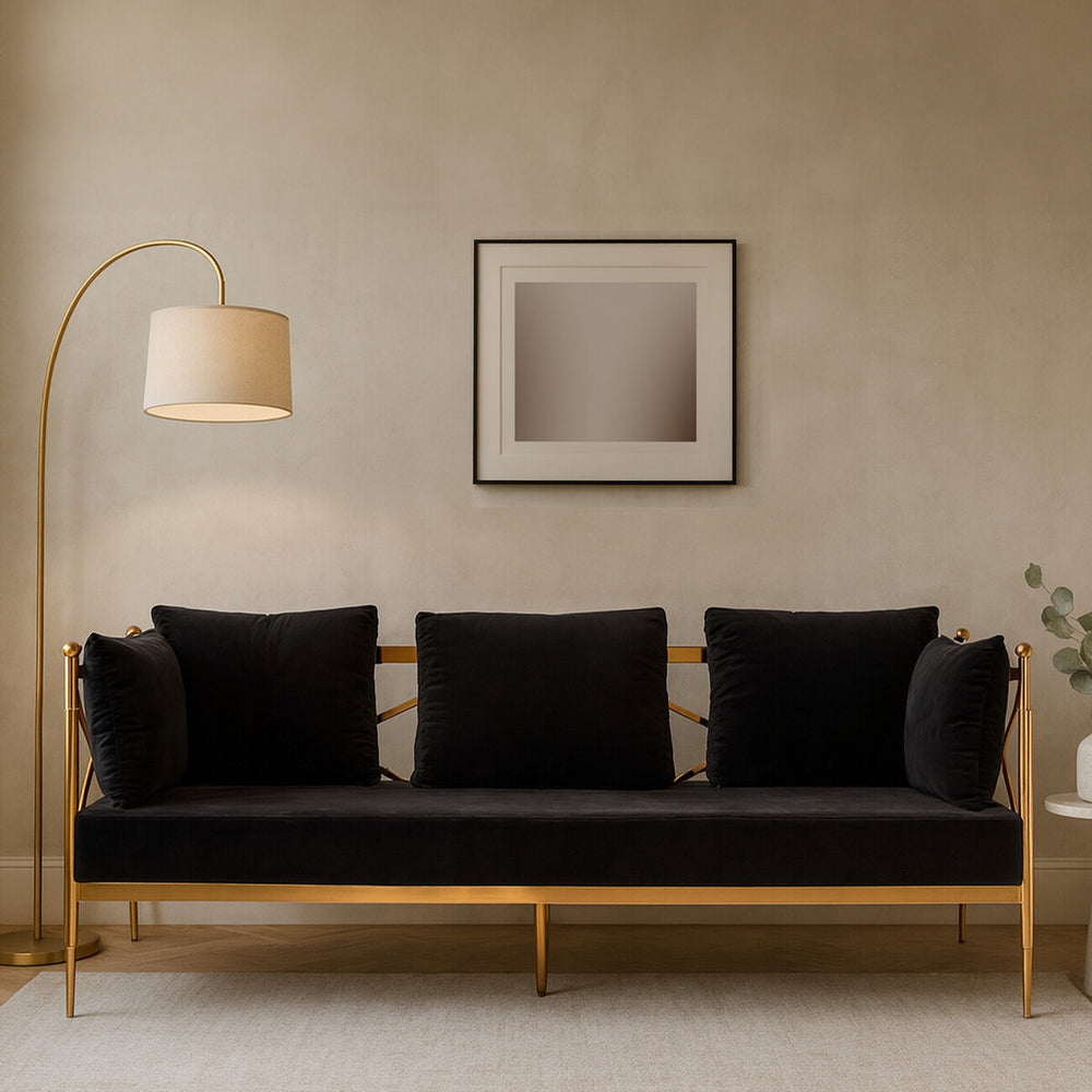 A sleek three-seater sofa upholstered in black velvet, featuring a stylish rose gold frame. The sofa has three plush cushions and is positioned against a neutral wall, complemented by a modern floor lamp and a minimalist artwork above it. A small potted plant is visible on the side, adding a touch of greenery to the setting