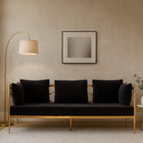 A sleek three-seater sofa upholstered in black velvet, featuring a stylish rose gold frame. The sofa has three plush cushions and is positioned against a neutral wall, complemented by a modern floor lamp and a minimalist artwork above it. A small potted plant is visible on the side, adding a touch of greenery to the setting