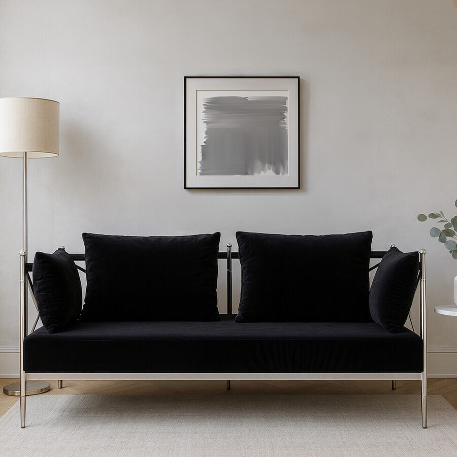 A sleek two-seater sofa upholstered in black velvet, featuring a modern silver frame. The sofa has two plush back cushions and a minimalist design, set against a neutral wall with a framed artwork above. A floor lamp is positioned to the left, adding to the contemporary aesthetic of the space