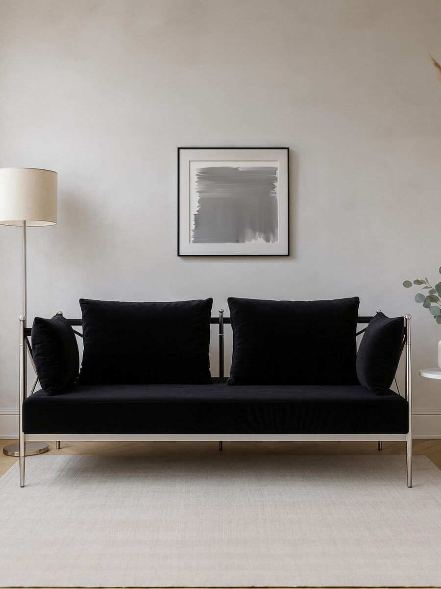 A sleek two-seater sofa upholstered in black velvet, featuring a modern silver frame. The sofa has two plush back cushions and a minimalist design, set against a neutral wall with a framed artwork above. A floor lamp is positioned to the left, adding to the contemporary aesthetic of the space
