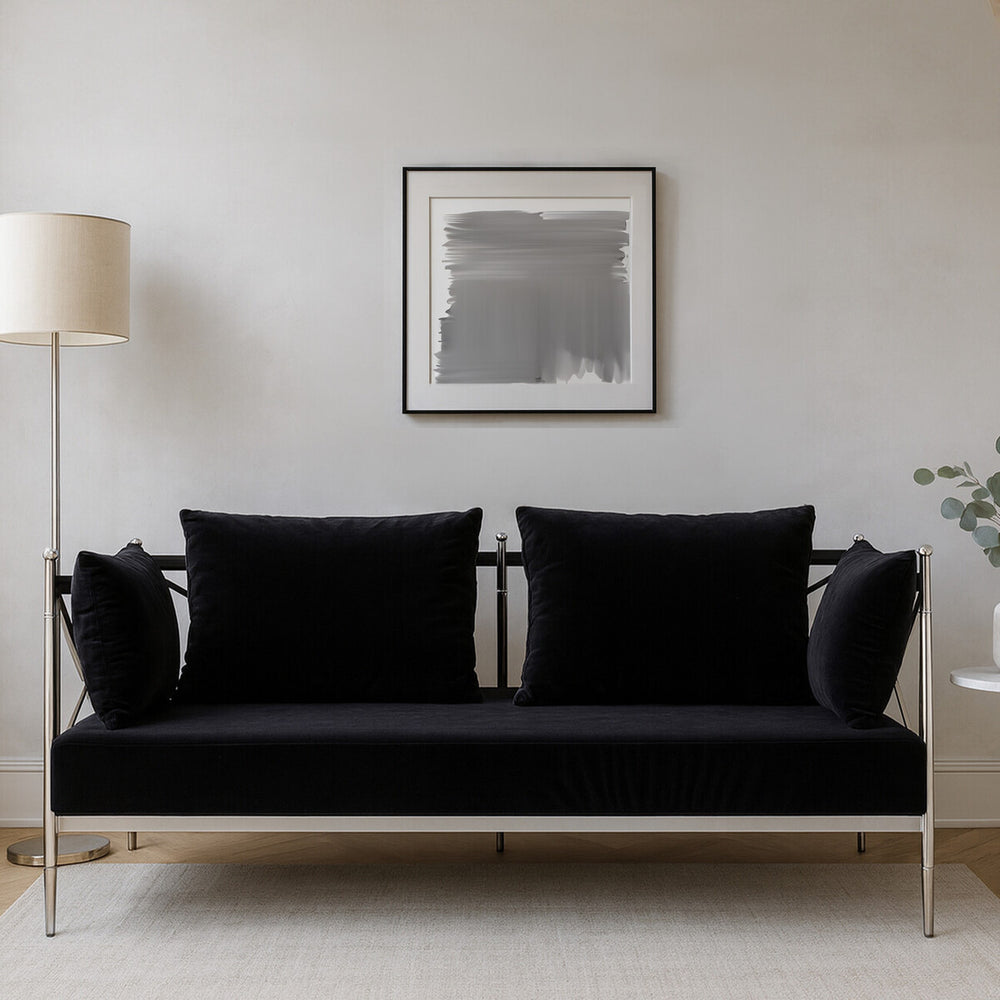 A sleek two-seater sofa upholstered in black velvet, featuring a modern silver frame. The sofa has two plush back cushions and is positioned against a neutral wall, with a floor lamp and a piece of abstract art in the background. A small plant is visible to the right of the sofa