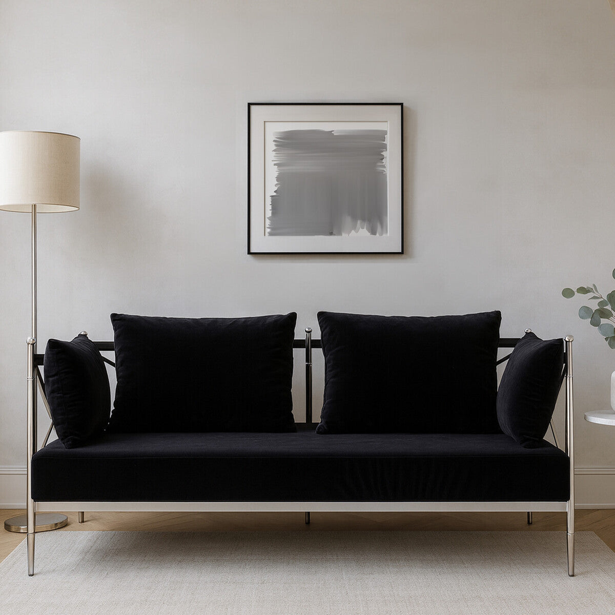 A sleek two-seater sofa upholstered in black velvet, featuring a modern silver frame. The sofa has two plush back cushions and is positioned against a neutral wall, with a floor lamp and a piece of abstract art in the background. A small plant is visible to the right of the sofa
