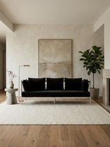 Black velvet three-seater sofa with a sleek silver frame, positioned against a light-colored wall. A large abstract artwork hangs above the sofa, and a potted plant is placed to the side. The floor features warm wooden tones, complemented by a light area rug