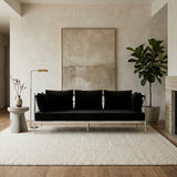 A sleek black velvet three-seater sofa with a silver frame, featuring three plush cushions, positioned in a modern living space with light-colored walls and wooden flooring. A large abstract artwork hangs on the wall behind the sofa, and a potted plant stands to the right, adding a touch of greenery to the room. A soft area rug lies beneath the sofa, enhancing the cozy atmosphere