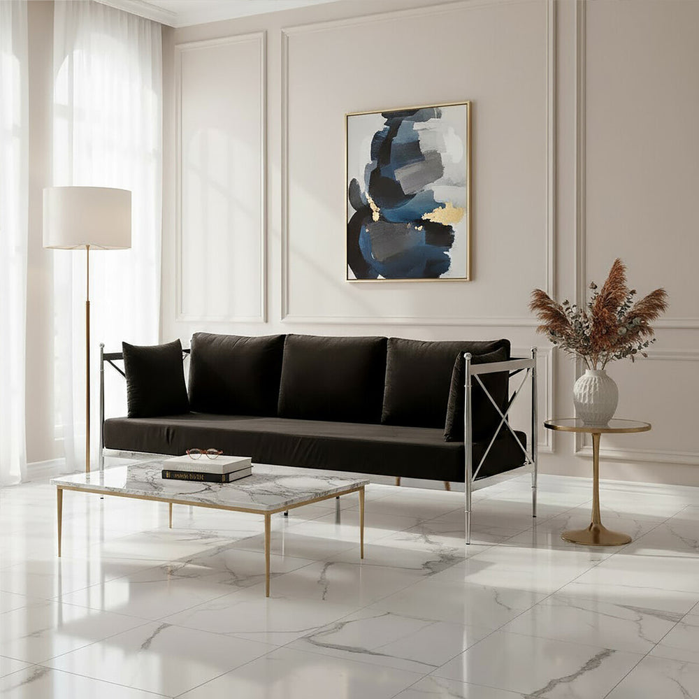 A sleek black velvet three-seater sofa with a modern silver frame, positioned in a bright room with large windows. The sofa features plush cushions and is complemented by a stylish glass coffee table and a decorative vase with dried flowers. A contemporary piece of artwork hangs on the wall behind the sofa, adding a touch of color to the neutral-toned space