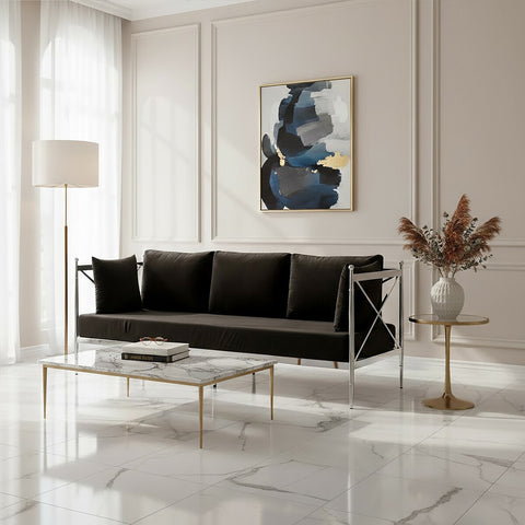 A sleek black velvet three-seater sofa with a modern silver frame, positioned in a bright room with large windows. The sofa features plush cushions and is complemented by a stylish glass coffee table and a decorative vase with dried flowers. A contemporary piece of artwork hangs on the wall behind the sofa, adding a touch of color to the neutral-toned space