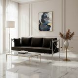 A sleek black velvet three-seater sofa with a modern silver frame, positioned in a bright room with large windows. The sofa features plush cushions and is complemented by a stylish glass coffee table and a decorative vase with dried flowers. A contemporary piece of artwork hangs on the wall behind the sofa, adding a touch of color to the neutral-toned space