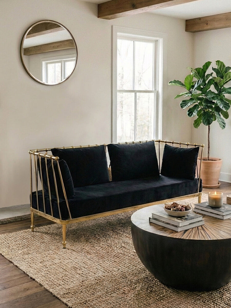 A stylish two-seater sofa featuring a black velvet upholstery and a sleek gold frame, positioned in a modern living room setting. The sofa has three plush cushions and is placed on a textured area rug, with a round coffee table in front. A large circular mirror hangs on the wall behind the sofa, and a potted plant adds a touch of greenery to the space. Natural light streams in through the