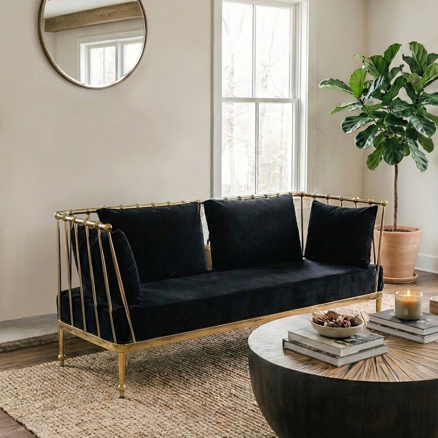 A stylish two-seater sofa featuring a black velvet upholstery and a sleek gold frame, positioned in a modern living room setting. The sofa has three plush cushions and is placed on a textured area rug, with a round coffee table in front. A large circular mirror hangs on the wall behind the sofa, and a potted plant adds a touch of greenery to the space. Natural light streams in through the