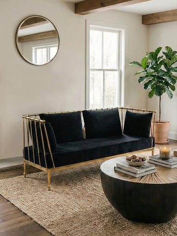 A stylish two-seater sofa featuring a black velvet upholstery and a sleek gold frame, positioned in a modern living room setting. The sofa has three plush cushions and is placed on a textured area rug, with a round coffee table in front. A large circular mirror hangs on the wall behind the sofa, and a potted plant adds a touch of greenery to the space. Natural light streams in through the