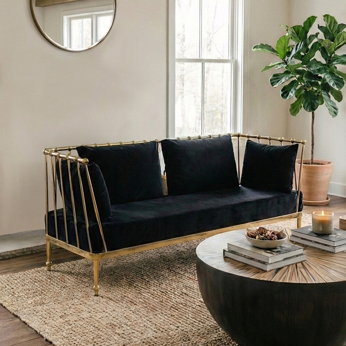 A stylish two-seater sofa featuring a black velvet upholstery and a sleek gold metal frame, positioned in a bright room with large windows. The sofa has three plush back cushions and is complemented by a round black coffee table and a potted plant nearby. A circular mirror hangs on the wall, reflecting the natural light in the space. The floor is covered with a textured area rug