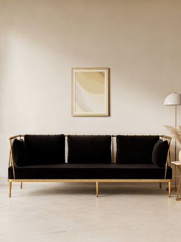 A sleek black velvet three-seater sofa with a gold finish base, featuring three plush cushions and a minimalist design, set against a neutral wall with a piece of abstract art and a modern floor lamp in the background