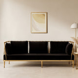 A sleek three-seater sofa upholstered in black velvet, featuring a gold finish frame. The sofa has three plush back cushions and a minimalist design, set against a neutral wall with a framed abstract artwork above. A modern floor lamp and a potted plant are visible in the background, enhancing the stylish living space