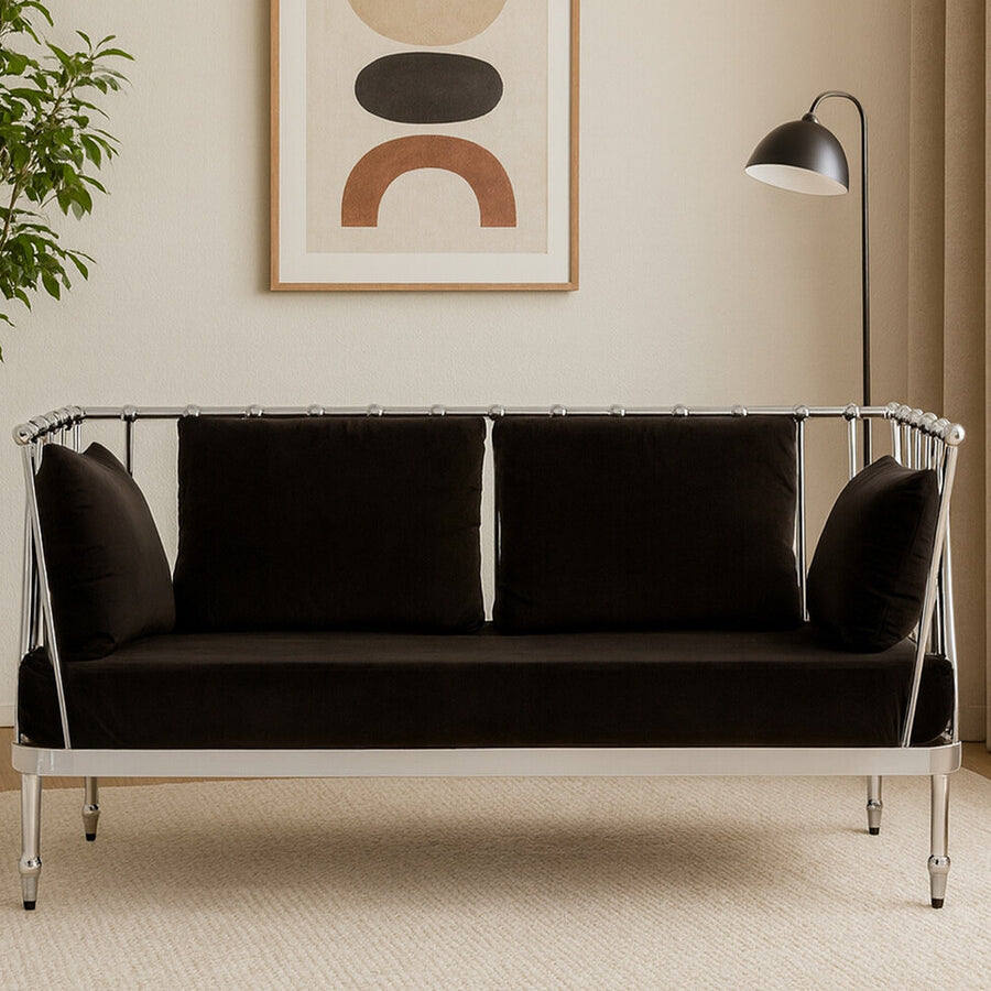 A stylish two-seater sofa featuring a sleek black velvet upholstery and a shiny silver frame, positioned in a modern living space with a decorative wall art piece and a floor lamp in the background