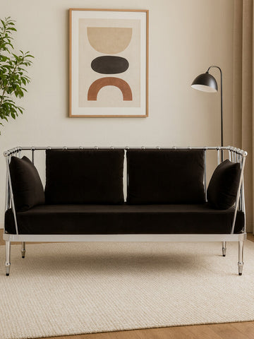 A stylish two-seater sofa featuring a sleek black velvet upholstery and a shiny silver frame, positioned in a modern living space with a decorative wall art piece and a floor lamp in the background