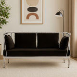 A sleek two-seater sofa upholstered in black velvet, featuring a modern silver metal frame. The sofa has plush back cushions and is positioned in a stylish interior setting with a floor lamp and abstract wall art in the background. The flooring is light-colored, complementing the overall aesthetic